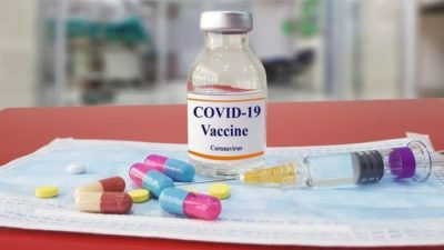 A glass jar labelled COVID_19 vaccine sits on a bench with capsules and precriptions drugs.