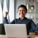 Smiling female investor holds hands up in victory in front of a laptop
