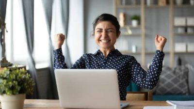 Smiling female investor holds hands up in victory in front of a laptop