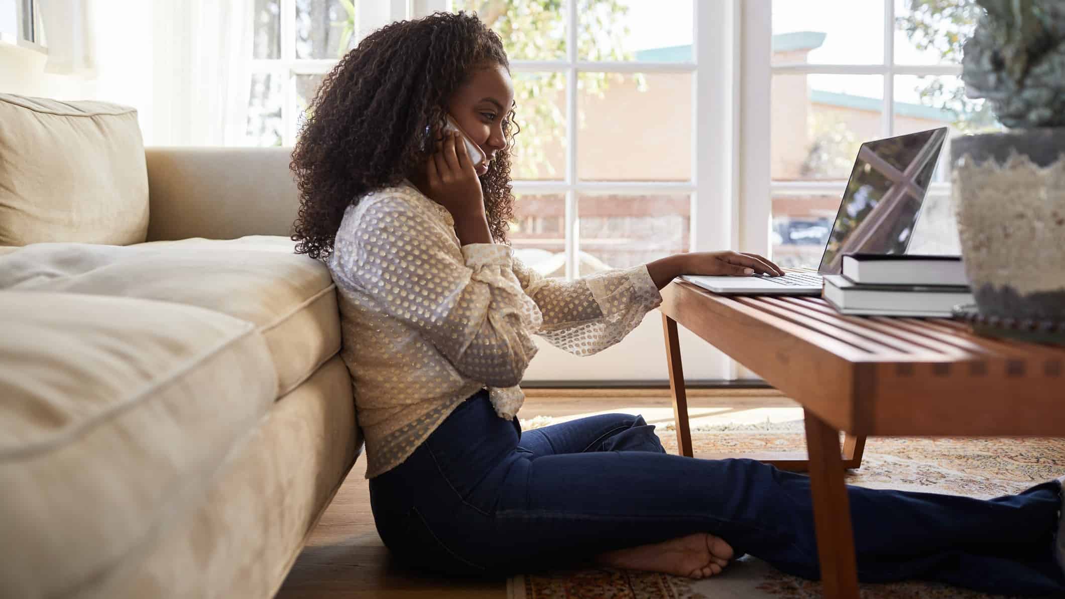 Young female investor smiling and speaking on mobile phone while sitting in front of laptop