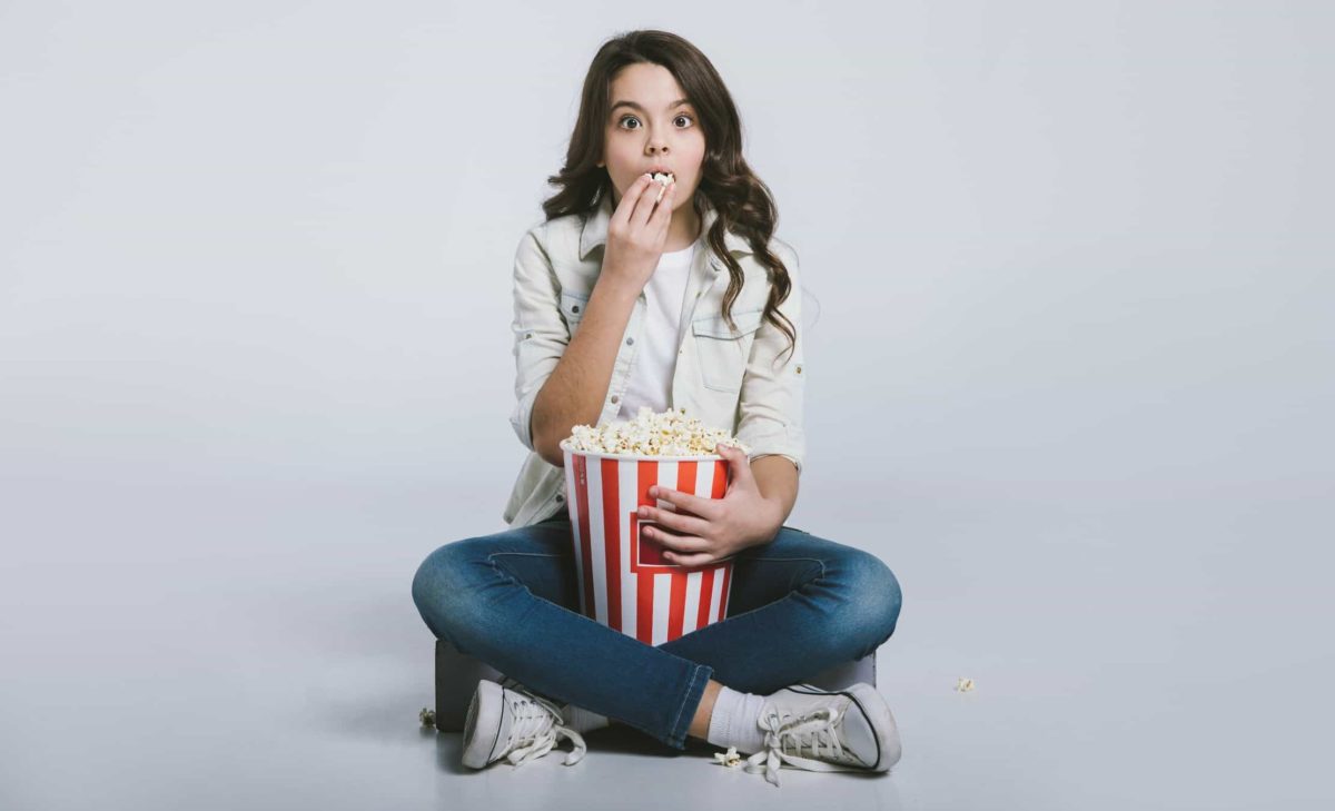 young woman sitting cross legged with large tub of popcorn and surprised facial expression