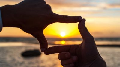 A sunset scene though the fingers of two hands, indicating the bigger picture