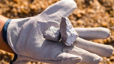 a gloved hand holds lumps of silver against a background of dirt as if at a mine site.