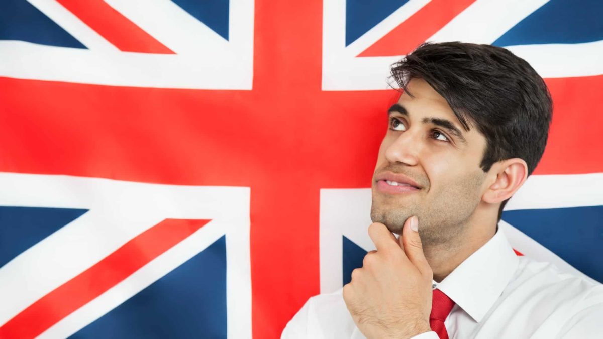 a man in a business shirt and tie holds his chin and ponders something in front of a backdrop of the british union jack flag
