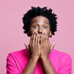 Scared, wide-eyed man in pink t-shirt with hands covering mouth.