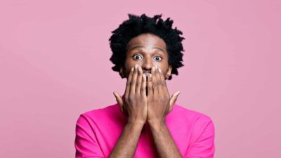 Scared, wide-eyed man in pink t-shirt with hands covering mouth