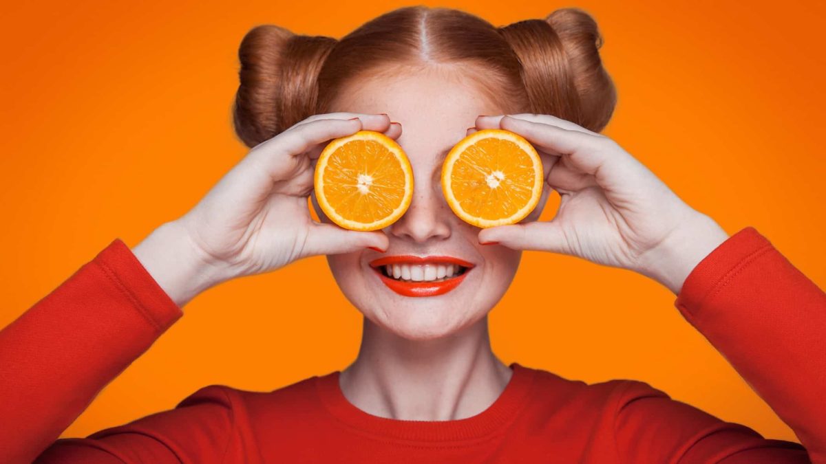 A smiling woman holds slices of orange to her eyes, indicating share price rises for ASX commodity shares