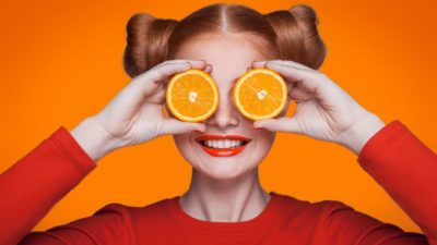 A smiling woman holds slices of orange to her eyes, indicating share price rises for ASX commodity shares