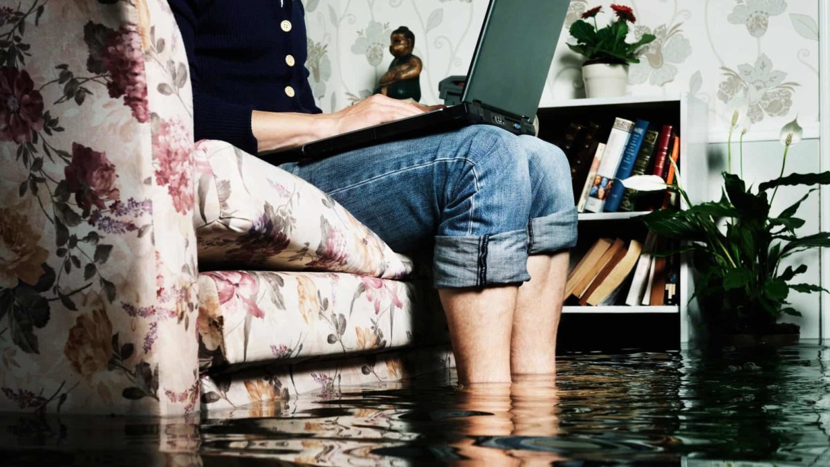 Person sitting on couch with computer on lap whilst flood waters rise around ankles