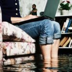Person sitting on couch with computer on lap whilst flood waters rise around ankles