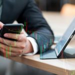 A share market investment manager monitors share price movements on his mobile phone and laptop