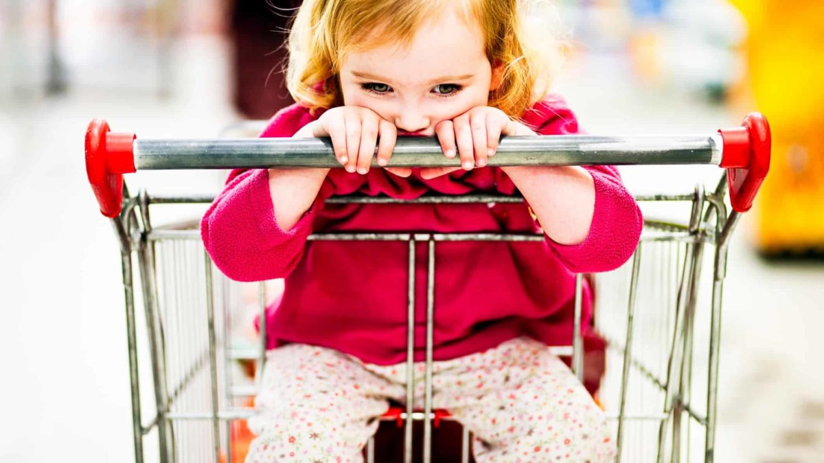 A sad little girl sits in a supermarket trolley, indicating a decline in share market price.