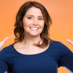 A smiling woman holds her hands up in front of an orange background.