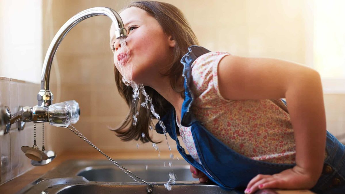 a young girl leans forward over a sink to take a drink of water directly from a running tap in a home setting.