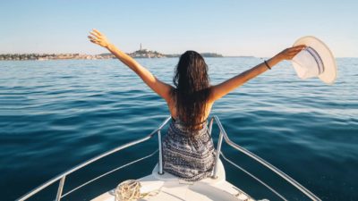 A young woman sitting atop a superyacht spreads her arms in joy, indictaing a share price rise for marine companies