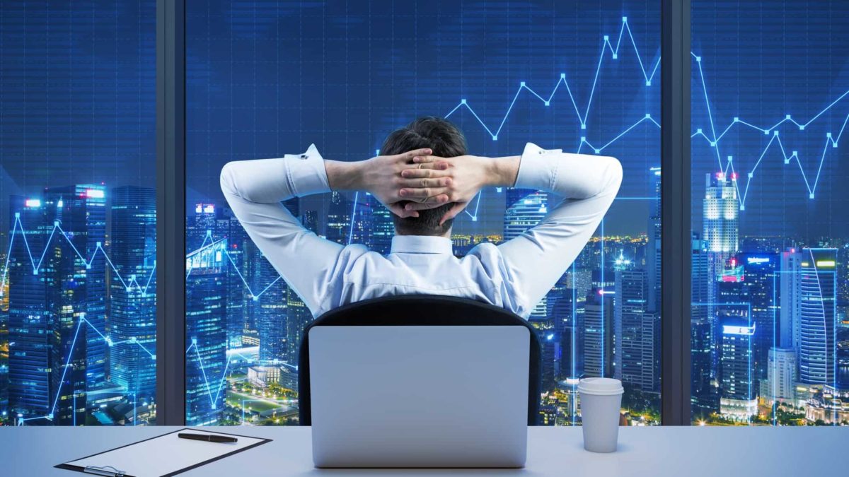 Business man at desk looking out window with his arms behind his head at a view of the city and stock trends overlay.