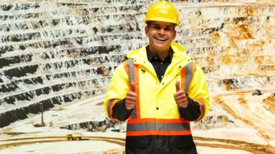 A smiling miner wearing a high vis vest and yellow hardhat and working for Superior Resources does the thumbs up in front of an open pit copper mine, indicating positive news for the company's share price today following a significant copper discovery