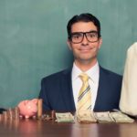 A smiling businessman sits at a desk with bags of mony, indicating a share price rise after funding has been approved