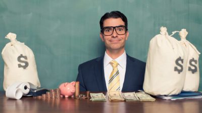 A smiling businessman sits at a desk with bags of mony, indicating a share price rise after funding has been approved