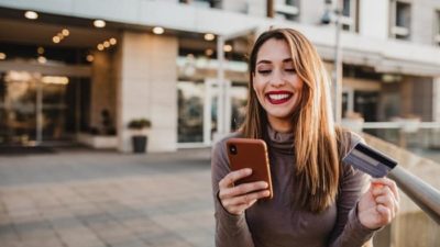 A happy woman stands outside a building looking at her phone and smiling widely
