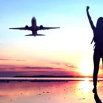 A woman stands on a runway with her arms outstretched in excitement with a plane in the air having taken off.