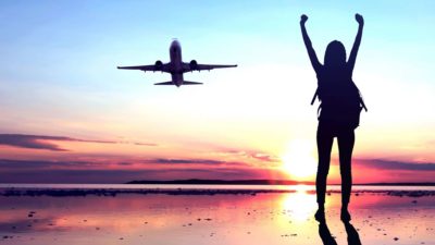 A woman stands on a runway with her arms outstretched in excitement as a plane takes off behind her representing the rising Qantas share price today