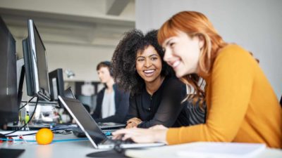 Two women happily smiling and working on their computers in an office