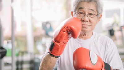 an elderly woman wearing boxing gloves raises one toward her face in a boxing pose while looking towards the camera with grey hair and spectacles on.