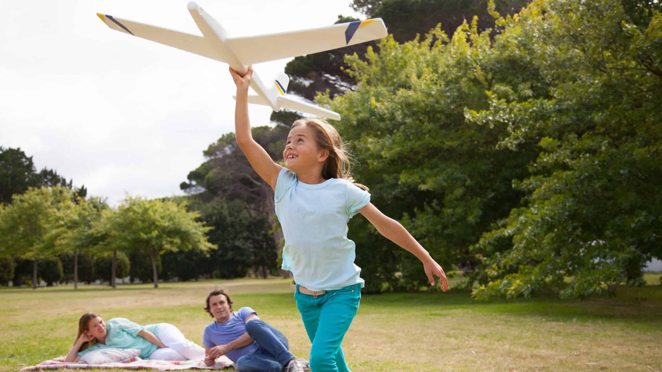 a girl runs with model plane in a park with her parents in the background lying on the grass watching her.