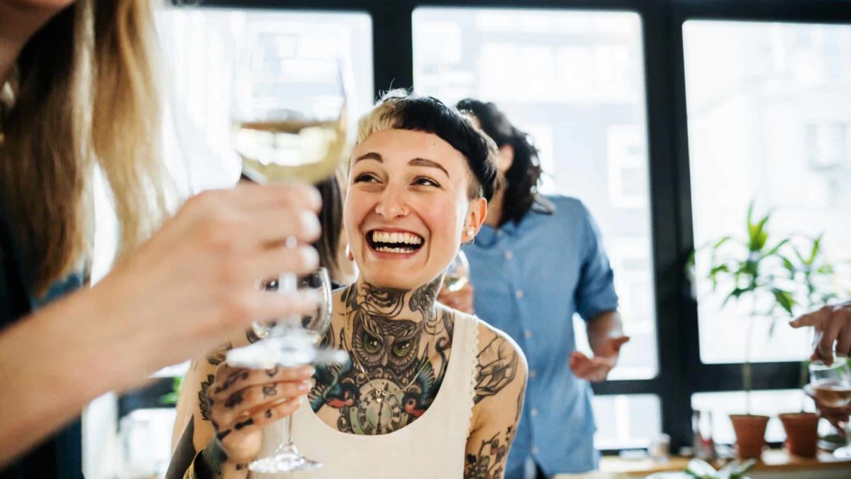 Smiling person with tattoos enjoying a glass of wine with a group of others.
