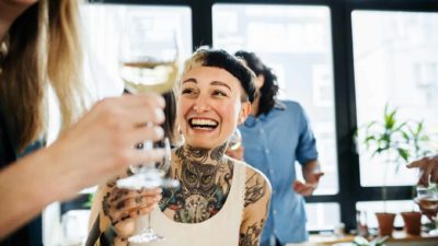 Smiling person with tattoos enjoying a glass of wine with a group of others.