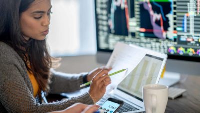 young woman reviewing financial reports at desk with multiple computer screens