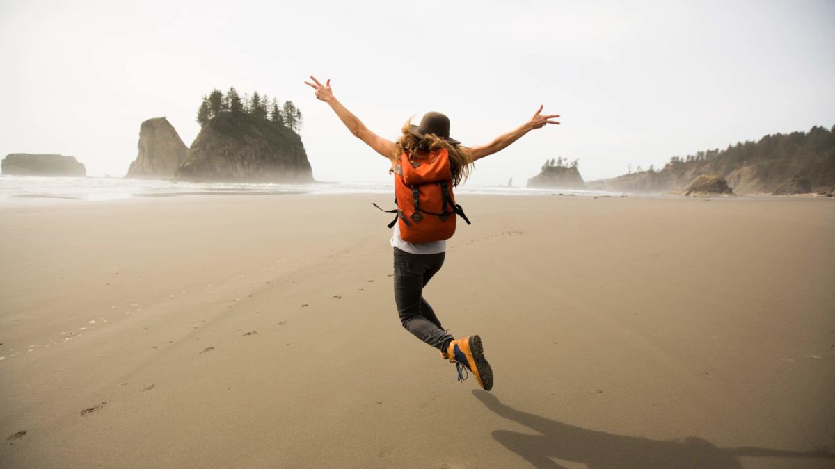 A woman wearing a backpack leaps for joy on the beach.
