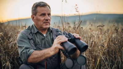 man sitting in field of grain with binoculars as if watching asx share price