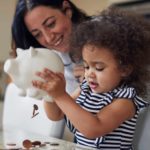 A young girl child empties coins out of her piggy bank with mum smiling over her shoulder.