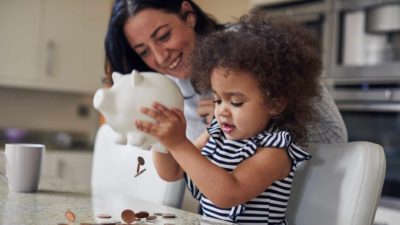 A young girl child empties coins out of her piggy bank with mum smiling over her shoulder.