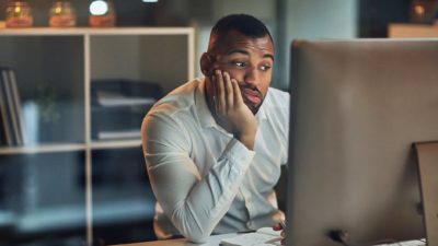 Bored man looking at his iMac with his head held in one hand feeling dismayed at AGL Energy's lower dividend