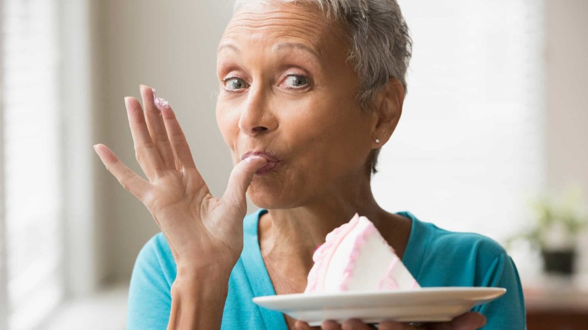 A mature woman holds a plate of cake and licks her thumb.