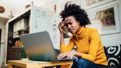 A woman sits on her lounge in front of her laptop looking concerned.