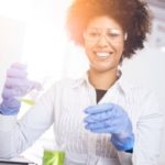 A medical researcher in a white coat holds laboratory equipment and smiles.