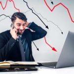 A shocked man sits at his desk looking at his laptop while talking on his mobile phone with declining arrows in the background representing falling ASX 200 shares today