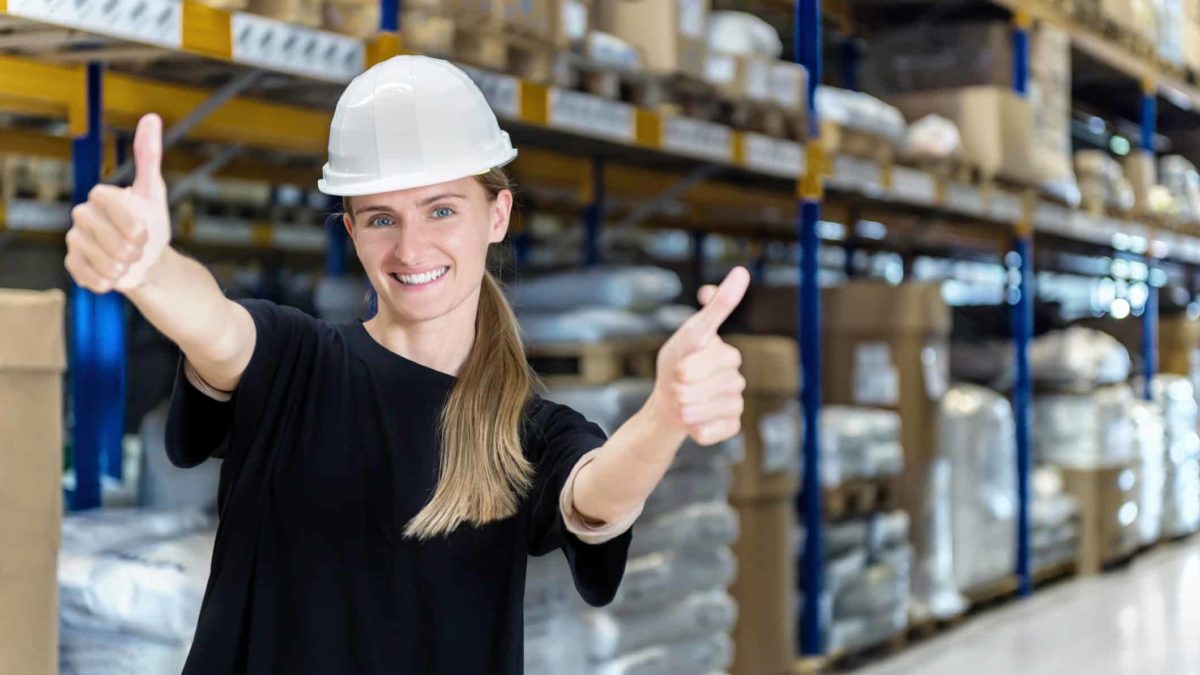 A happy woman in a hard hat gives two thumbs up, standing in a packing warehouse.