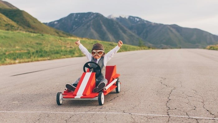 kid riding a plastic go kart with his hands raised in the air with mountains in the background symbolising winning a race