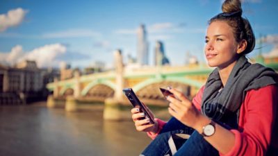 A smiling young woman sits on a bridge in London checking her online shopping, indicating share price movement for ASX BNPL shares overseas