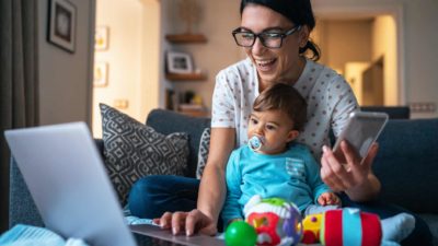 A smiling woman sits in front of her laptop with her baby in her lap looking at her rising ASX shares including the VAS ETF