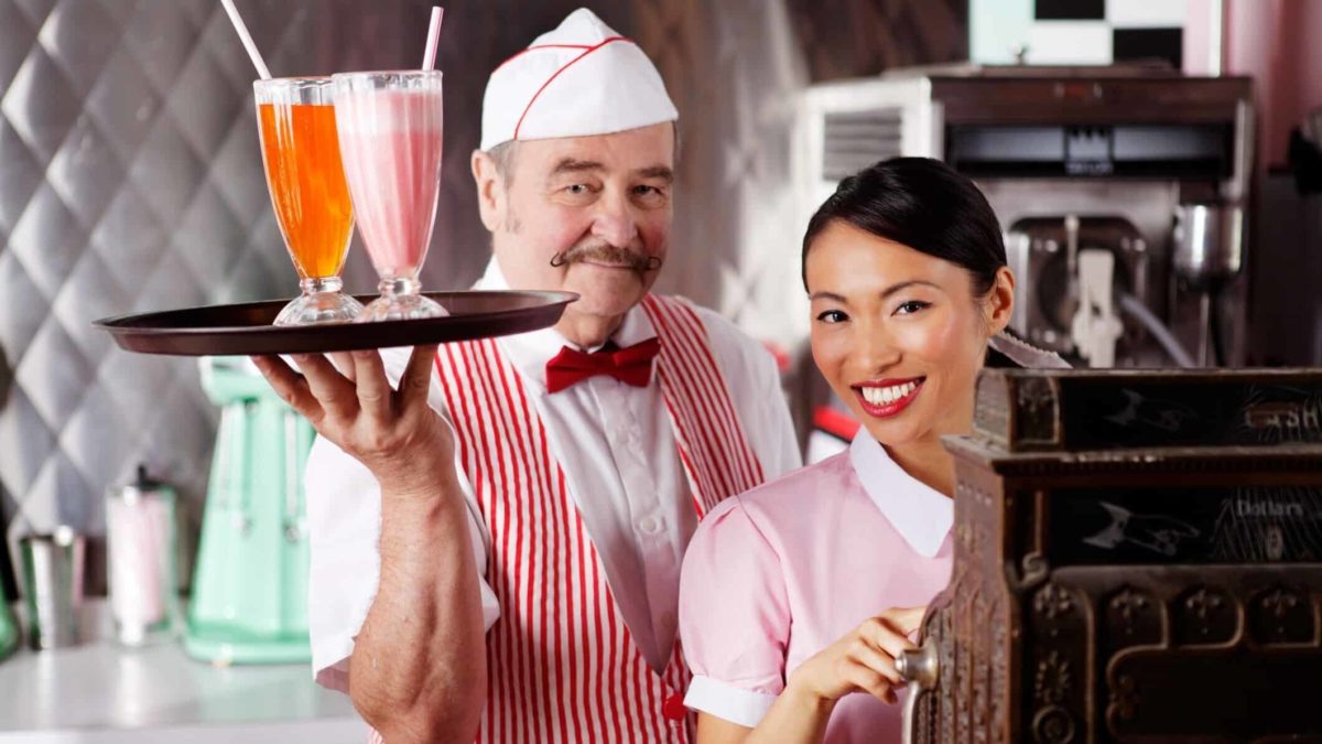 An older gentleman waiter dressed in a red and white 1950s-style diner uniform holding a tray of milkshakes and a young Asian waitress in the same uniform standing next to a traditional cash register