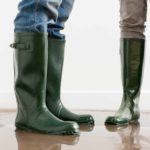 Legs and feet of two people wearing green gumboots standing in a flooded room ready to clean up.
