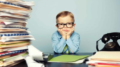 a child dressed as businessperson looking sad and dejected at desk with pile of papers and old fashioned telephone.