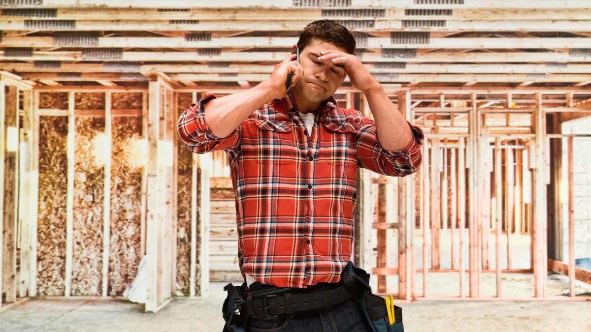 Sad Probuild construction worker in front of half built house puts his hand to his forehead as he talks on the phone