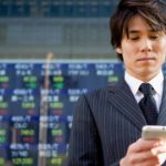 stock market news, person checks phone in front of electronic stock exchange boad
